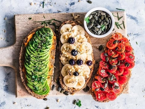 A cutting board with three pieces of toast topped with avocado, banana, and tomato